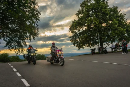 Bayern © Moppetfoto.de Zwei Motorradfahrer fahren bei Sonnenuntergang an einer Kurve vorbei.