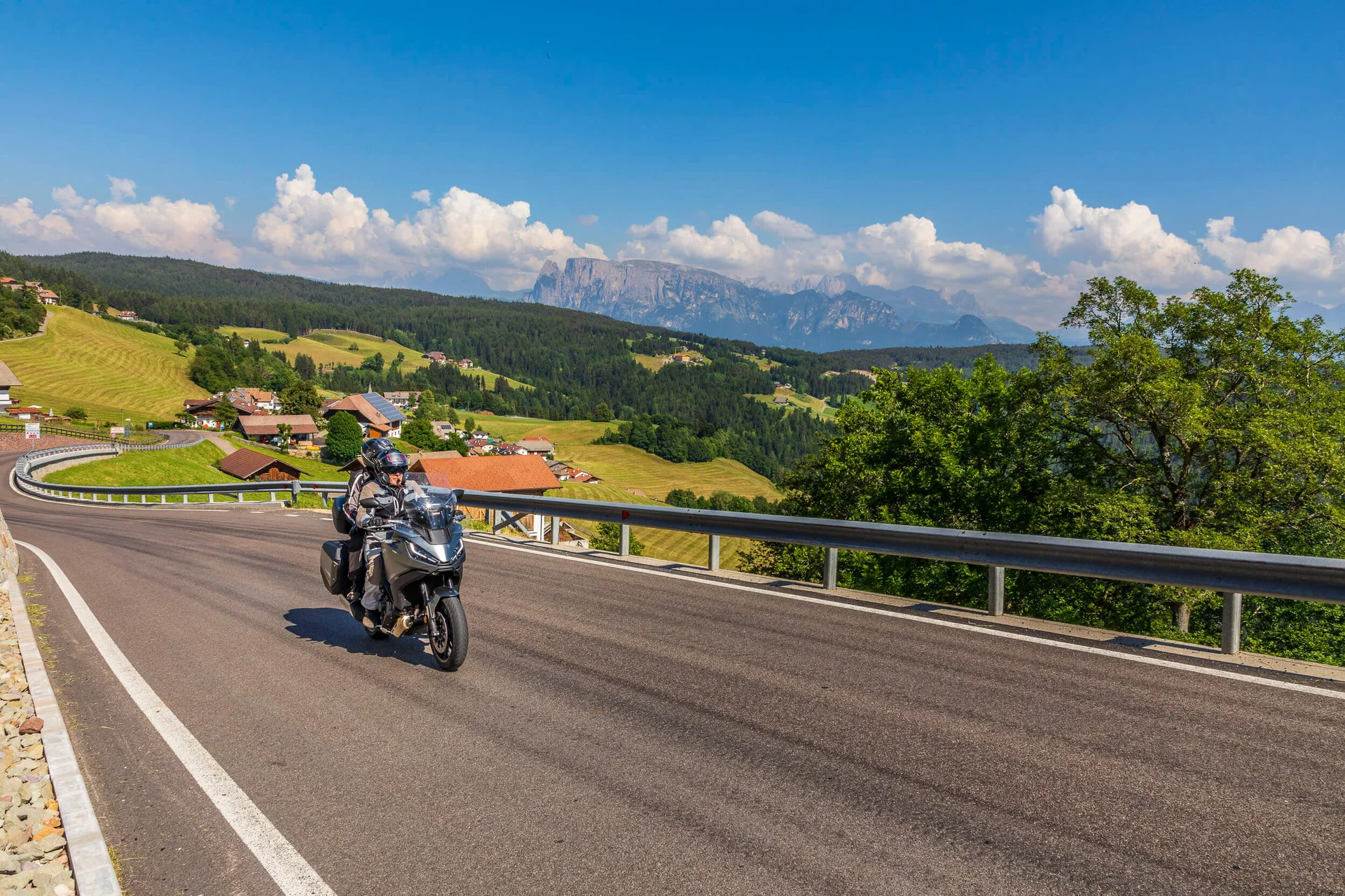 Motorvakantie © Alexander Seger Motorrijders op kronkelende bergweg met dorp en bergen op de achtergrond