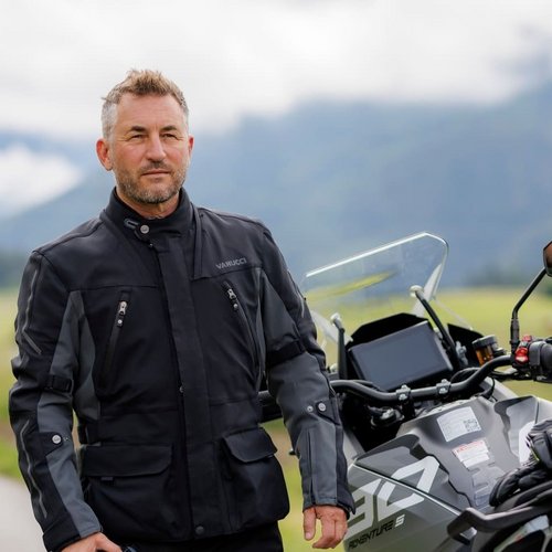 Motorradurlaub © Detlev Louis Motorrad-Vertriebsgesellschaft mbh Mann in schwarzer Motorradkleidung hält Helm neben Motorrad auf Landstraße