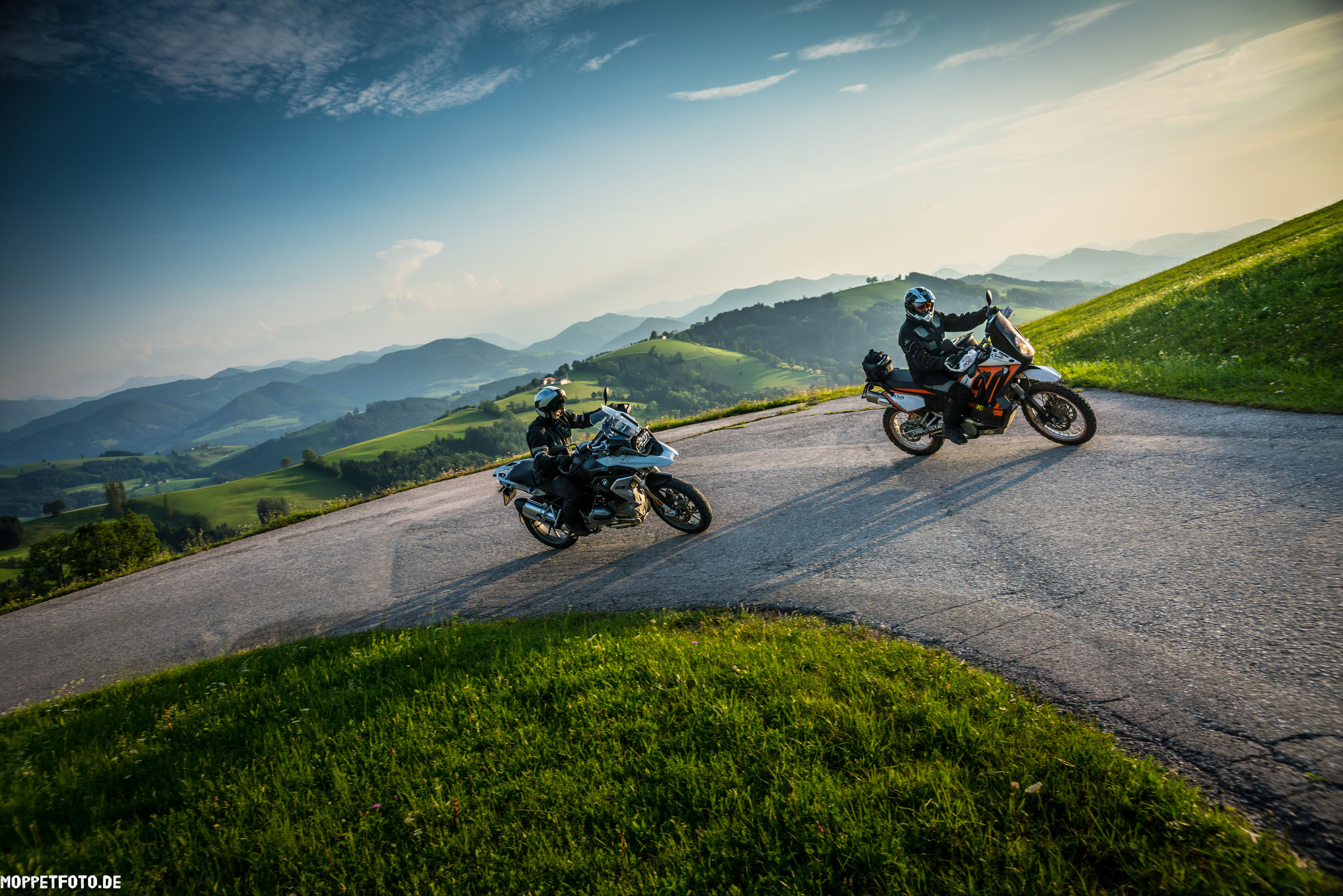 Motorradurlaub © Moppetfoto.de Zwei Motorradfahrer auf kurviger Bergstraße mit grünen Hügeln und blauem Himmel