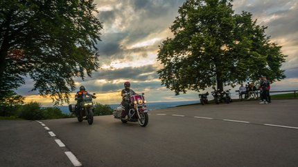 Bayern © Moppetfoto.de Zwei Motorradfahrer fahren bei Sonnenuntergang an einer Kurve vorbei.