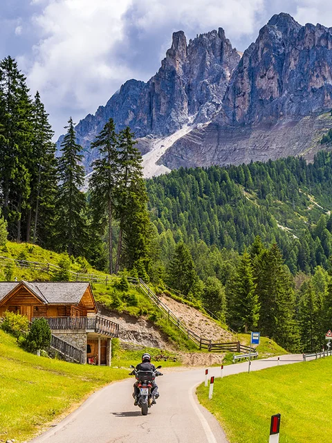 Motorradurlaub © Alexander Seger Motorradfahrer auf kurviger Straße in den Bergen mit Holzhütte und Nadelbäumen