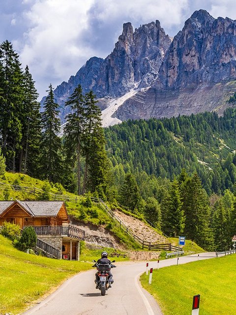 Motorradurlaub © Alexander Seger Motorradfahrer auf kurviger Straße in den Bergen mit Holzhütte und Nadelbäumen