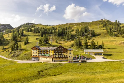 Almhotel & Genussgasthof Hierzegger © Alexander Seger Hotel in grüner Berglandschaft mit Sessellift und Traktor im Vordergrund