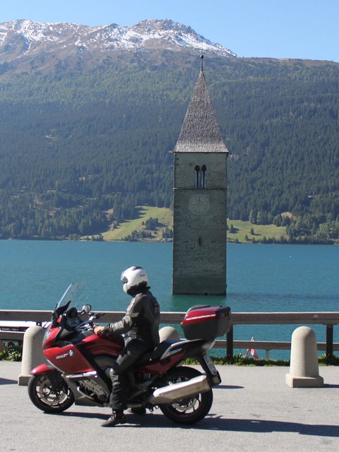 Motorradurlaub © The Dilitz Company GmbH Motorradfahrer vor versunkenem Kirchturm im See mit Bergen im Hintergrund