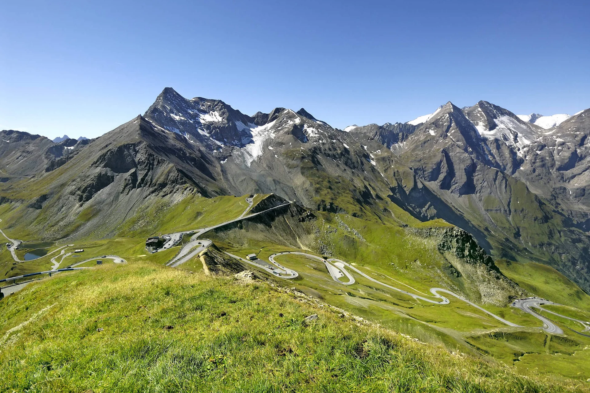 Motorradurlaub © Großglockner Hochalpenstraßen AG Serpentinenstraße in grünen Alpen mit schneebedeckten Bergen unter blauem Himmel