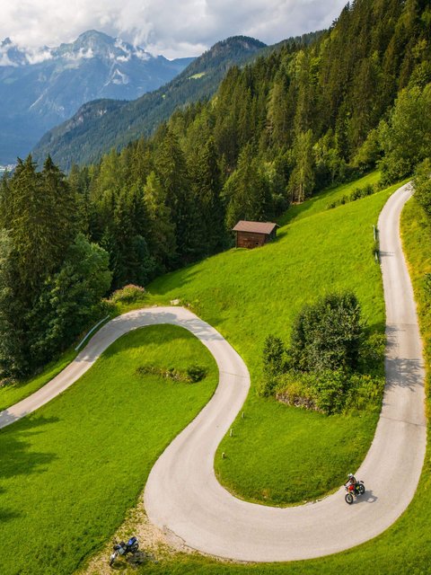 Motorradurlaub © Moppetfoto.de Kurvige Bergstraße mit Motorradfahrern und Wald vor Alpenkulisse