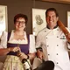 Motorradurlaub © The Dilitz Company GmbH Frau mit Weinflasche und Mann in Kochjacke mit Pfanne und Nudelholz