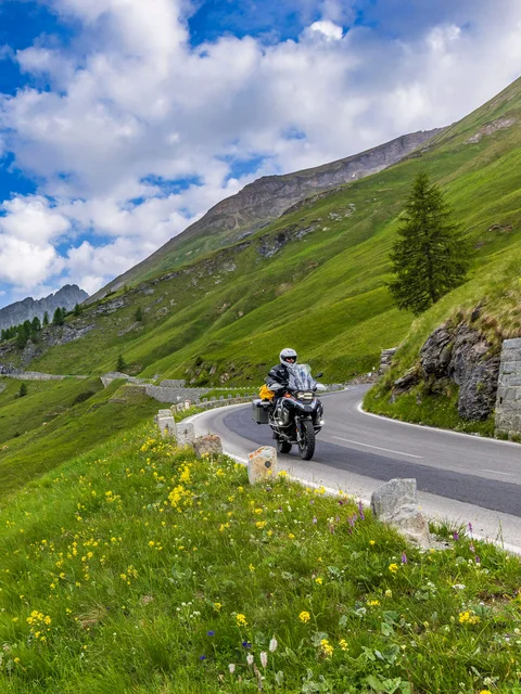 Motorradurlaub © Alexander Seger Motorradfahrer auf kurvenreicher Bergstraße mit grünen Wiesen und schneebedeckten Gipfeln